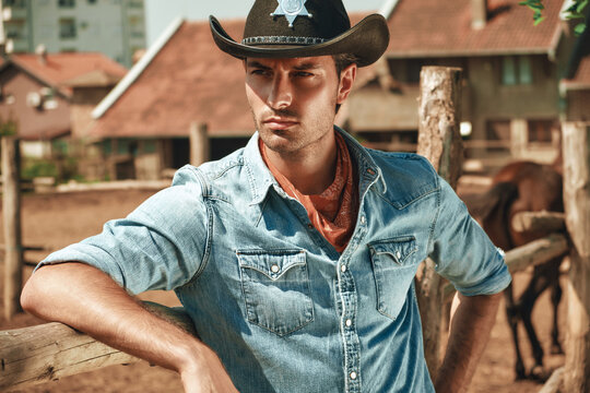 Young man dressed as a cowboy leans against a wooden fence in a countryside location. Horses graze nearby, while rustic buildings create a charming backdrop during sunny weather