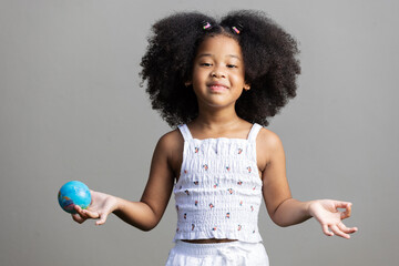 A confident young girl smiles while holding a small globe in one hand, standing against a plain background. Her pose and expression convey joy, curiosity, and a sense of global awareness.