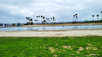 Distant view at Santa Clara Point in Sail Bay across a patch of green grass lawn, beach sand and narrow stretch of water under cloudy morning sky, San Diego, California