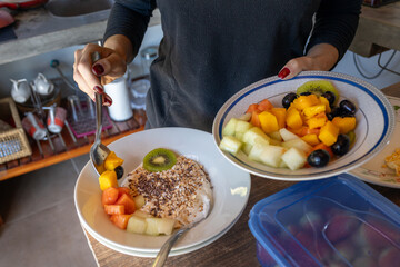 woman preparing a bowl with yogurt mango in a healthy breakfast with cereal and honey, grapes, kiwi, banana, mango and papaya