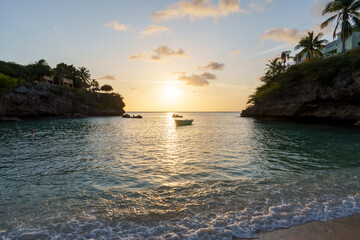 Naklejka premium Pôr do sol em enseada tropical de Curaçao, com águas calmas, barcos ao longe e céu dourado, transmitindo serenidade, beleza natural e atmosfera caribenha.