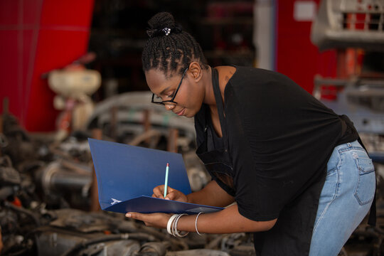 Smiling african american female worker holding blue folder while inspecting car parts warehouse. Concept of automotive maintenance, quality control service. Employees checking inventory.