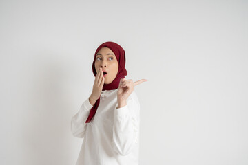 Young woman wearing a red hijab and white top expressing surprise while pointing sideways. Studio shot against a white background, highlighting emotions, cultural attire, and a moment of astonishment.