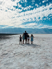 A father walks hand-in-hand with four children across the salt flats of Badwater Basin under a dramatic desert sky.
