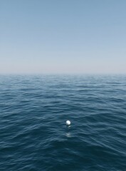 A solitary white sphere floats gently on a vast expanse of calm, dark blue ocean, beneath a clear, pale blue sky