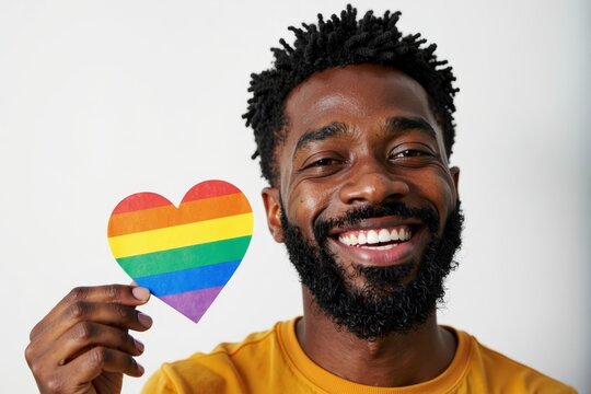 Happy African American man with a clean-shaven beard proudly clutching a rainbow heart on a white backdrop, representing support and acceptance within the LGBT community.