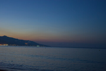 A coastal city's lights illuminate the shoreline at dusk with a dark silhouette of mountains in the background.