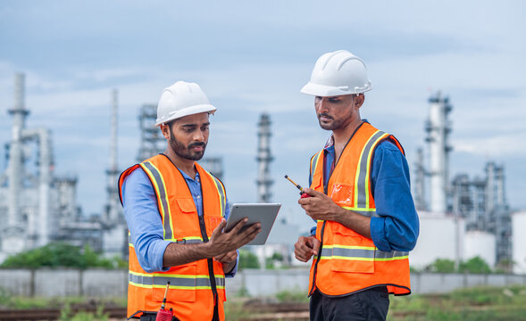 Engineers wearing safety gear, including hard hats examining survey tablet standing industrial facility gas task or oil refinery engaged in a job requires high safety standards concept.	