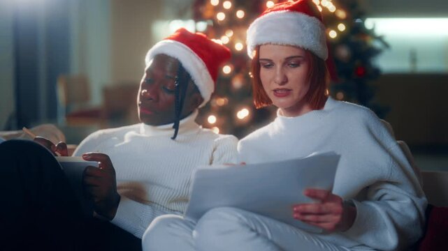 Christmas time, portrait of black man and caucasian woman making gift list. Young people preparing to merry Xmas party at night, writing shopping list, dreaming about future, happy loving couple