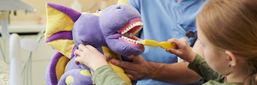 Caucasian girl child brushing teeth of large plush dragon toy held by adult male dentist, demonstrating proper dental hygiene technique during educational session at dental clinic - Powered by Adobe