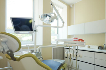 Modern dental office showing empty dental chair with overhead lamp, monitor, dental instruments and model teeth on counter, bright clean environment with large windows