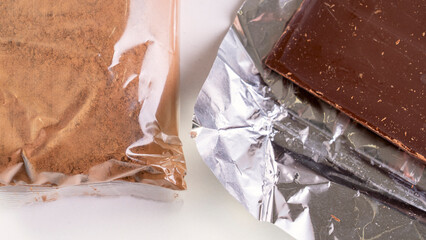 A bag of chocolate powder is next to a foil wrapper of chocolate. The foil wrapper is partially torn, and the chocolate is partially exposed