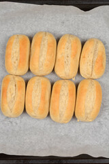 Eight freshly baked, oval-shaped rolls are arranged in two rows on baking paper, photographed from above
