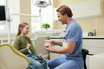 Obraz premium Caucasian male dentist showing dental model to Caucasian girl child sitting in dental chair, both smiling and interacting in modern dental clinic with equipment visible