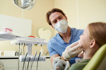 Caucasian young adult male dentist examining mouth of Caucasian child girl in dental clinic, dentist wearing mask and gloves while child holding stuffed animal during dental checkup