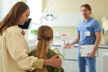 Caucasian woman comforting Caucasian child while listening to male doctor in medical uniform holding clipboard, standing in modern clinic examination room