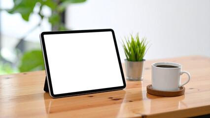 A digital tablet device with a blank screen for mockup, placed on a wooden office desk with a coffee cup and small plant.