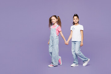 Obraz premium Full body side profile view little two friends kids children girls 9-10 years old wearing casual clothes together hold hands walk go look camera isolated on plain purple background. Lifestyle concept.