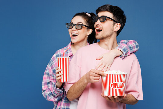 Young excited couple two friends family man woman they wear pink casual clothes together 3d glasses watch movie film hold bucket of popcorn cup of soda pop in cinema isolated on plain blue background.