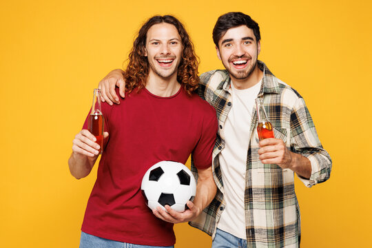Young two friends men sport fans wearing casual clothes cheer up support football team hold soccer ball watch tv live stream drink beer hug look camera isolated on yellow background studio portrait.