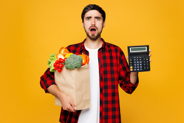 Young sad mad man wear casual clothes hold craft paper bag for takeaway mock up with food products show calculator device isolated on plain yellow orange background Delivery service from shop concept