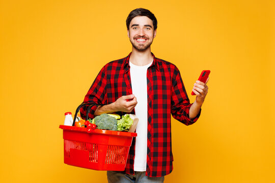 Young smiling happy man wear casual clothes hold basket bag for takeaway mock up with food products use mobile cell phone isolated on plain yellow orange background Delivery service from shop concept