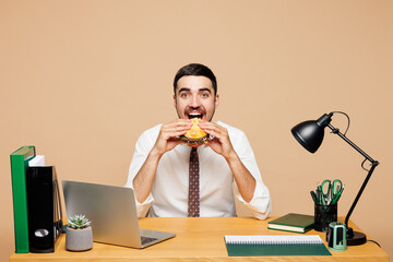 Young successful hungry employee business man wears white shirt tie sit work at office desk with pc laptop eat biting fast food burger isolated on plain beige background. Achievement career concept.