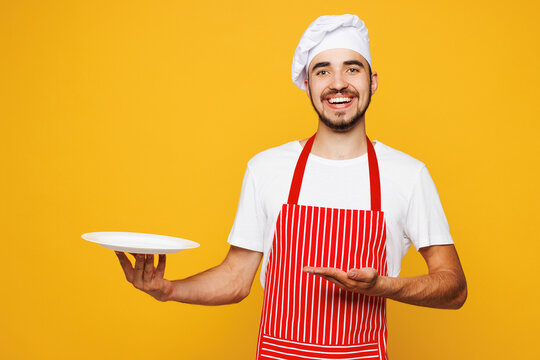 Young smiling fun housewife housekeeper chef cook baker man wear red apron toque hat hold in hand point on empty plate isolated on plain yellow orange background studio portrait. Cooking food concept.