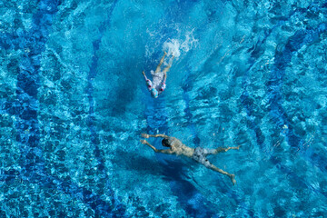 Top-down view of two swimmers enjoying a bright blue swimming pool, with sunlight creating shimmering patterns on the water surface.