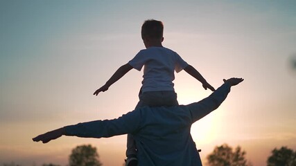 Boy lifts arms out riding on shoulders. Silhouette of child and father in sunset glow. Outdoor freedom in evening light. Kid on shoulders with open arms. Father supports boy in golden field.