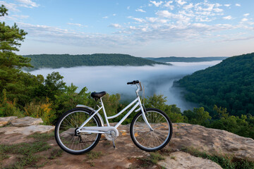 Fototapeta premium White Bicycle on Rock Overlooking Foggy Mountain Landscape at Sunrise or Sunset