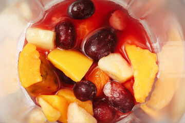 Fresh fruit pieces in blender. Top view of mango, pineapple, cherries and grapes with red juice. Ingredients prepared for smoothie or juice. Healthy natural drink raw ripe tropical and berry fruits.
