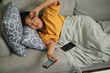 High angle view shot, Middle-aged Asian woman is lying on a sofa, looking drowsy while watching a movie in the living room at home.