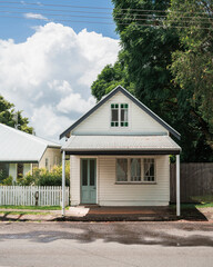 Small white wooden house with a green door, corrugated metal roof, and covered porch, set against lush trees and a bright, cloudy sky, Dunoon, Australia. © pauline.mongarny