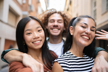 Fototapeta premium Three cheerful young multiracial friends hugging and taking selfie portrait in the city