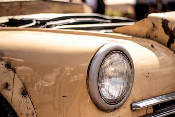 Fototapeta premium Detailed view of an old beige car’s front headlight and hood, showing rust spots and visible damage in bright sunlight.