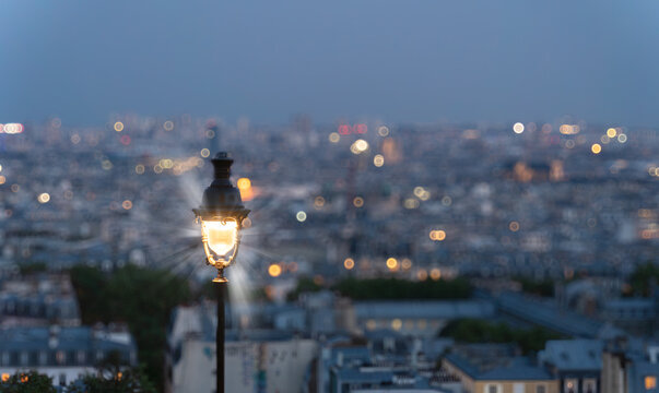 Fototapeta Parisian street lamp glows warmly against a blurred backdrop of the city lights at dusk, creating a romantic and nostalgic atmosphere