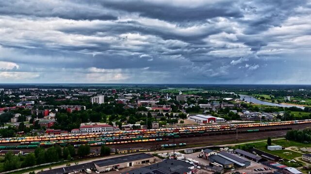 Aerial view of Jelgava&rsquo;s main industrial train station in Latvia, with long rows of colorful freight cars in the foreground, the cityscape and the Lielupe River stretching beyond.