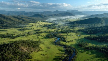Obraz premium Aerial view of a valley with winding river, lush green fields, and distant mountains, shrouded in morning mist