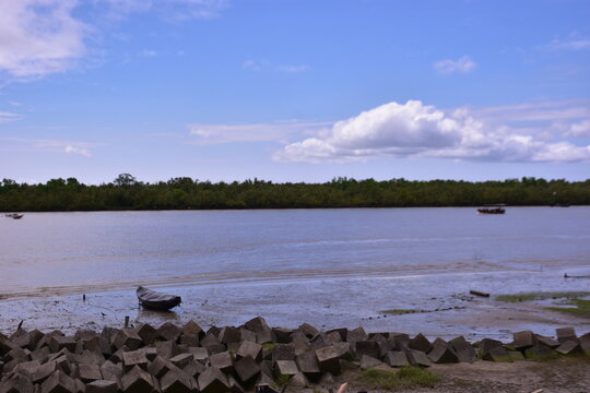 A scenic view of the Bakkhali River estuary at low tide in Bangladesh, showing muddy tidal flats, a concrete seawall, and fishing boats. - Powered by Adobe
