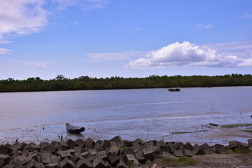 A scenic view of the Bakkhali River estuary at low tide in Bangladesh, showing muddy tidal flats, a concrete seawall, and fishing boats.