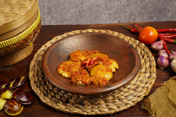 Close-up of spicy Indonesian jengkol balado served in a traditional clay plate, surrounded by fresh chili, tomato, and shallots on rustic background.