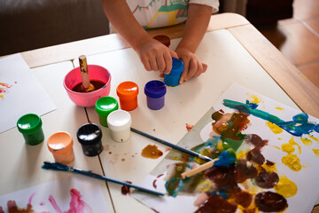 A young child opens a paint jar during painting activity at home with daylight. Various paint pots...