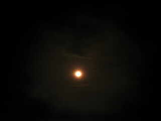 A bright, glowing full moon is captured in the night sky, surrounded by a faint halo and partially veiled by thin, dark clouds