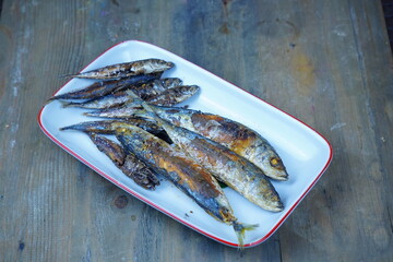 A rectangular white plate with a red rim holds several fried sardines on a weathered wooden table.