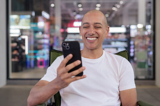 Happy bald Latino man sitting outdoors in coffee shop smiling and using mobile phone