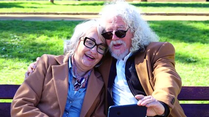 Happy senior couple sitting close together on a park bench, smiling as they take a selfie with a smartphone. Both wear stylish jackets and sunglasses, enjoying a sunny day outdoors in a relaxed - Powered by Adobe