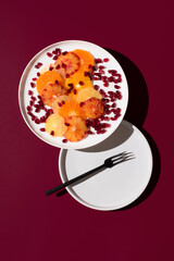 Rodajas de cítricos variados: naranja, limón, mandarina, naranja sanguina, lima y semillas de granada en un plato blanco sobre fondo granate. Vista superior