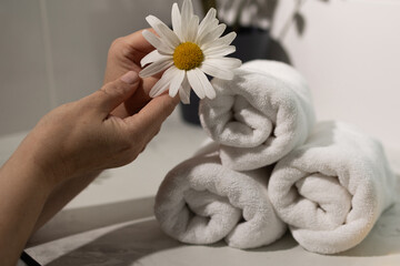 Soft white towels are arranged neatly while a gentle hand holds a daisy above them, creating a calming atmosphere in a modern bathroom. The scene evokes tranquility and care.