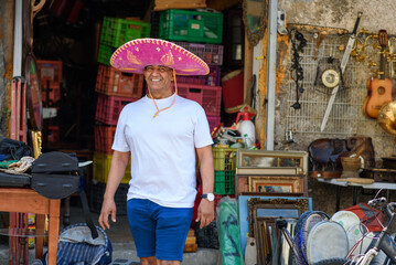 A cheerful man tourist wearing a vibrant sombrero walks outside a bustling market shop. The background features a variety of colorful items, suggesting a lively atmosphere.